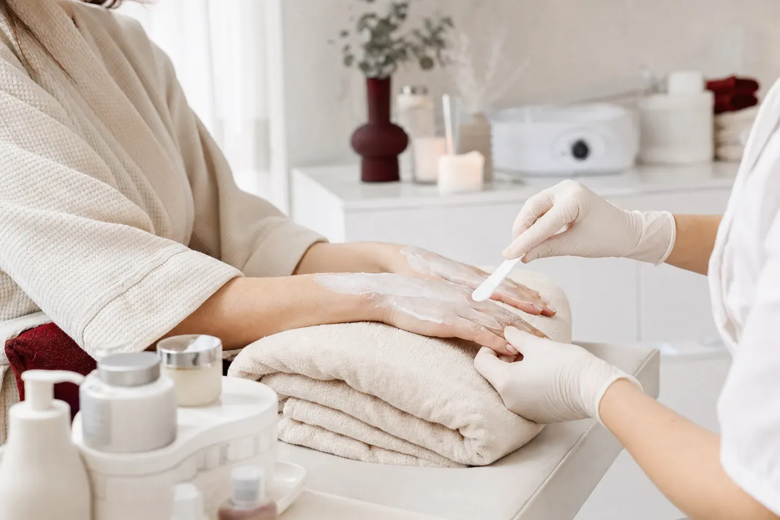 A cosmetologist applies paraffin cream to a client’s hands resting on a towel