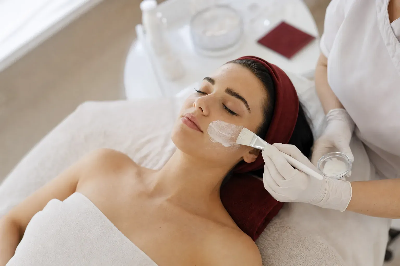 A cosmetologist brushes a cleansing mask onto a young woman’s cheek